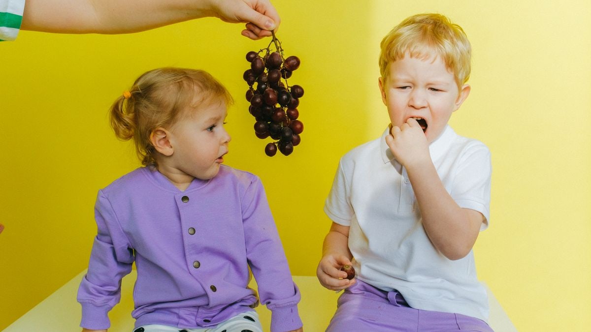 A boy enjoying grapes while sitting next to his curious sister indoors against a yellow background.